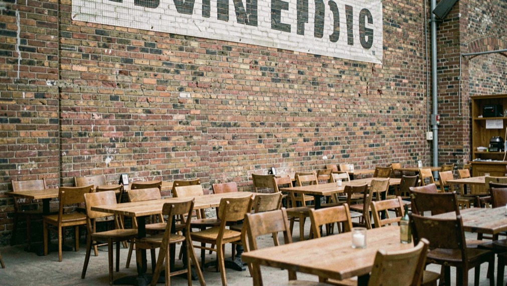 Breed scherm hoog aan een bakstenen muur, daaronder rijen tafels met stoelen schuin naar voren gedraaid.