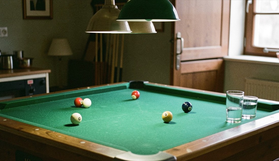 Biljartlaken in groen vilt onder een hanglamp, keu op de band, halfvolle glazen op een rand.