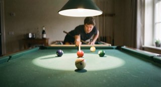 Groen biljartlaken met drie ballen en een keu in aanslag, hanglamp werpt een cirkel licht op het laken.