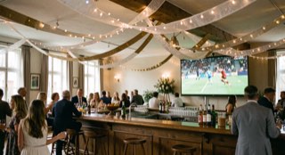 Feestelijk verlichte zaal met slingers boven de bar en een scherm dat een wedstrijd toont.