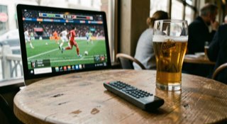 Afstandsbediening op een cafétafel voor een scherm met een lopende wedstrijd, glas bier ernaast.