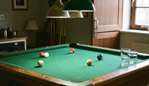Biljartlaken in groen vilt onder een hanglamp, keu op de band, halfvolle glazen op een rand.